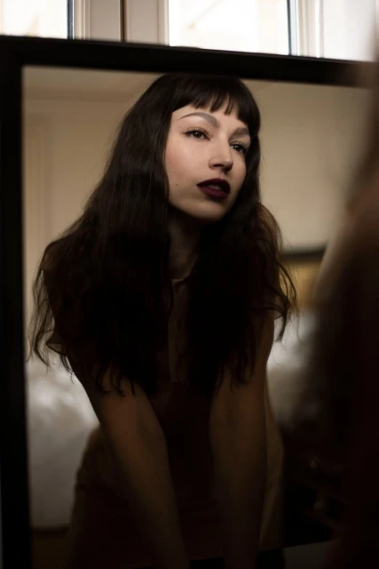 Portrait of a brunette woman with dark lipstick reflected in a mirror indoors, with a neutral color palette and soft lighting.