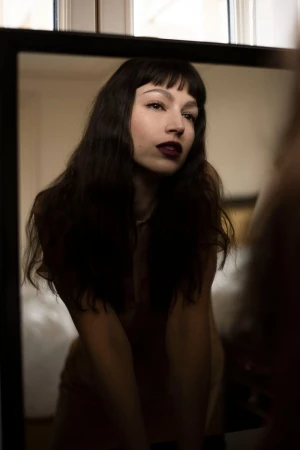 Portrait of a brunette woman with dark lipstick reflected in a mirror indoors, with a neutral color palette and soft lighting.