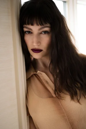 Close-up portrait of Úrsula Corberó with dark hair and bangs leaning against a wall, wearing a tan collared shirt.