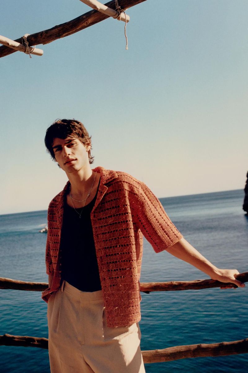 Male model in open knit shirt poses on a wooden structure overlooking the ocean under a clear blue sky in an outdoor fashion shot.