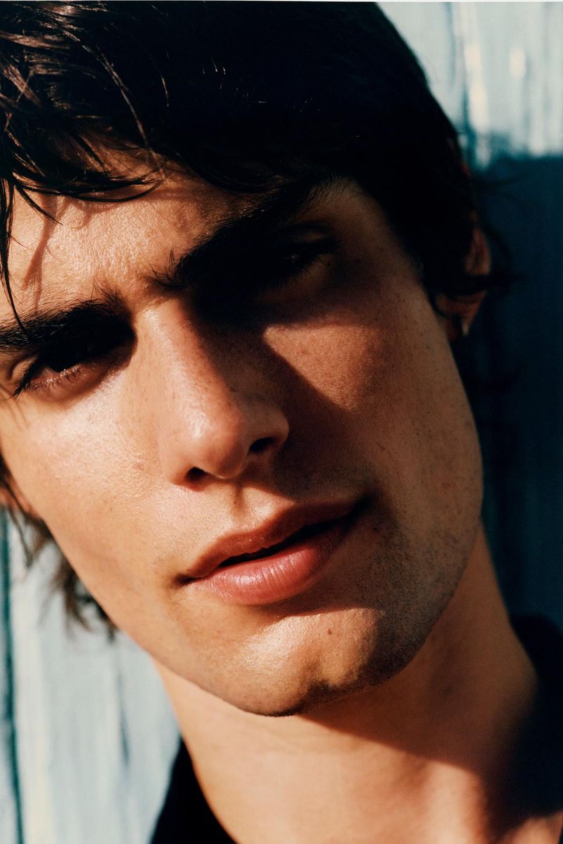 Portrait of a young adult male with dark hair and light skin, wearing a dark top, lit by bright, natural light against a blue wall.