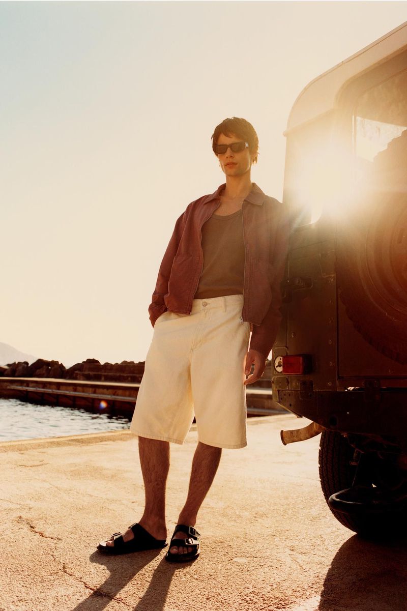 Man in jacket, shorts, and sandals stands by car on waterfront in golden light. Fashion editorial portrait with outdoor setting.