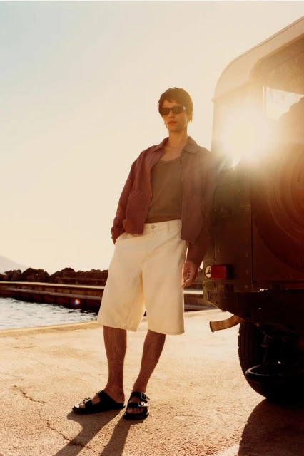 Man in jacket, shorts, and sandals stands by car on waterfront in golden light. Fashion editorial portrait with outdoor setting.