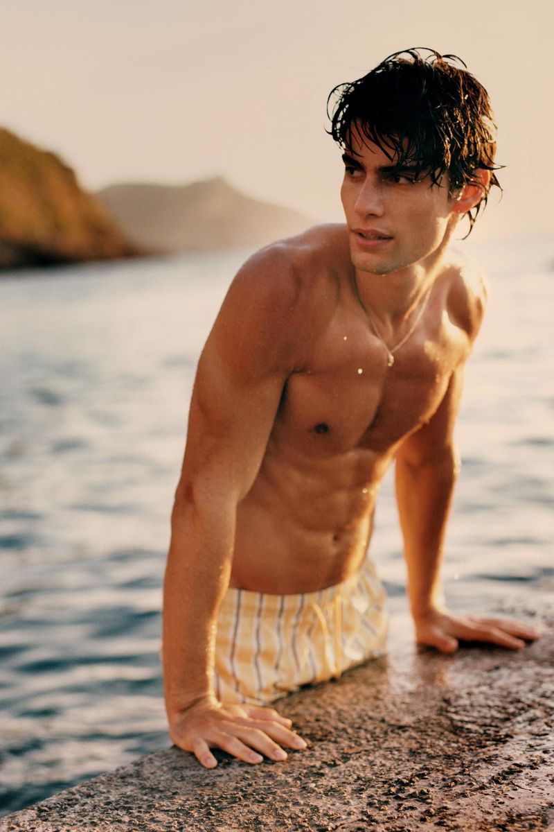 A shirtless, muscular adult male poses at the edge of the water, wearing plaid boxer shorts, with a shoreline and headland in the background.