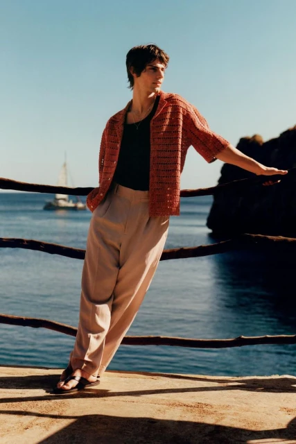 Person leaning against a wooden rail with a view of the sea and a boat, wearing orange shirt and beige pants in outdoor daylight.
