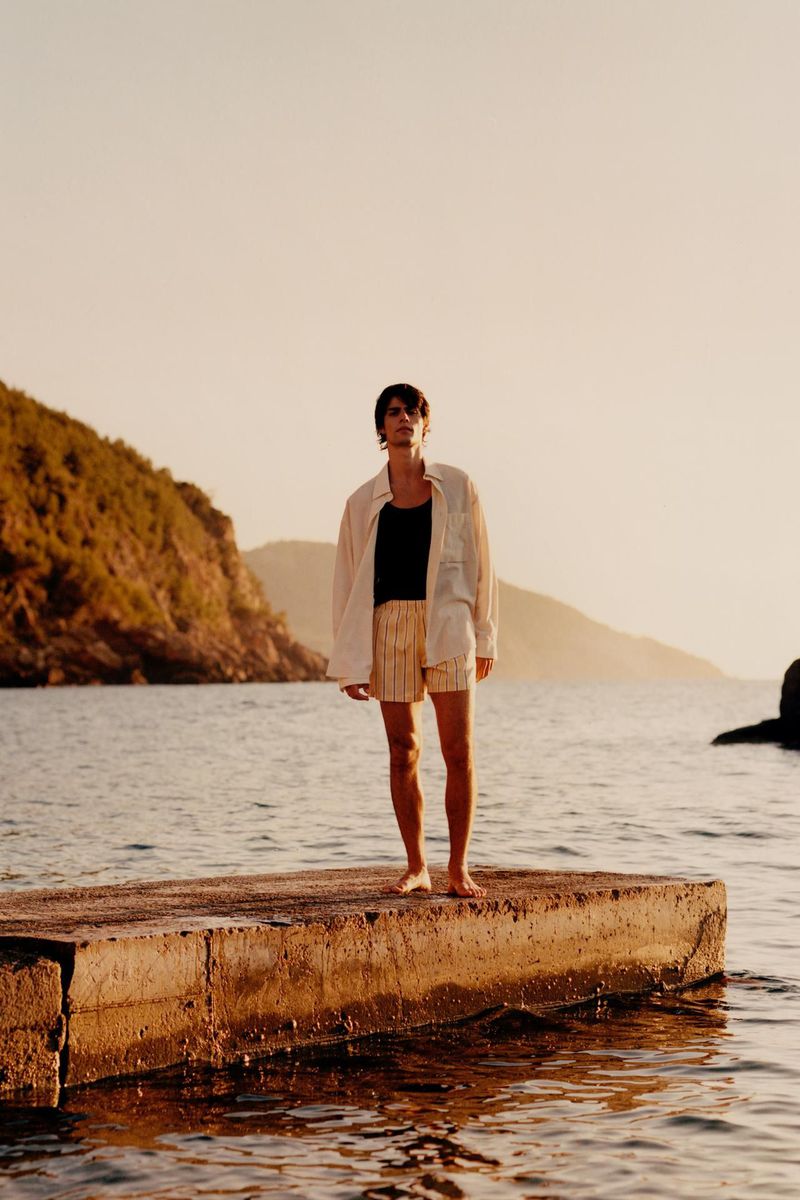 Woman in casual summer wear stands barefoot on a pier overlooking the water at sunset with lush vegetation and mountains.