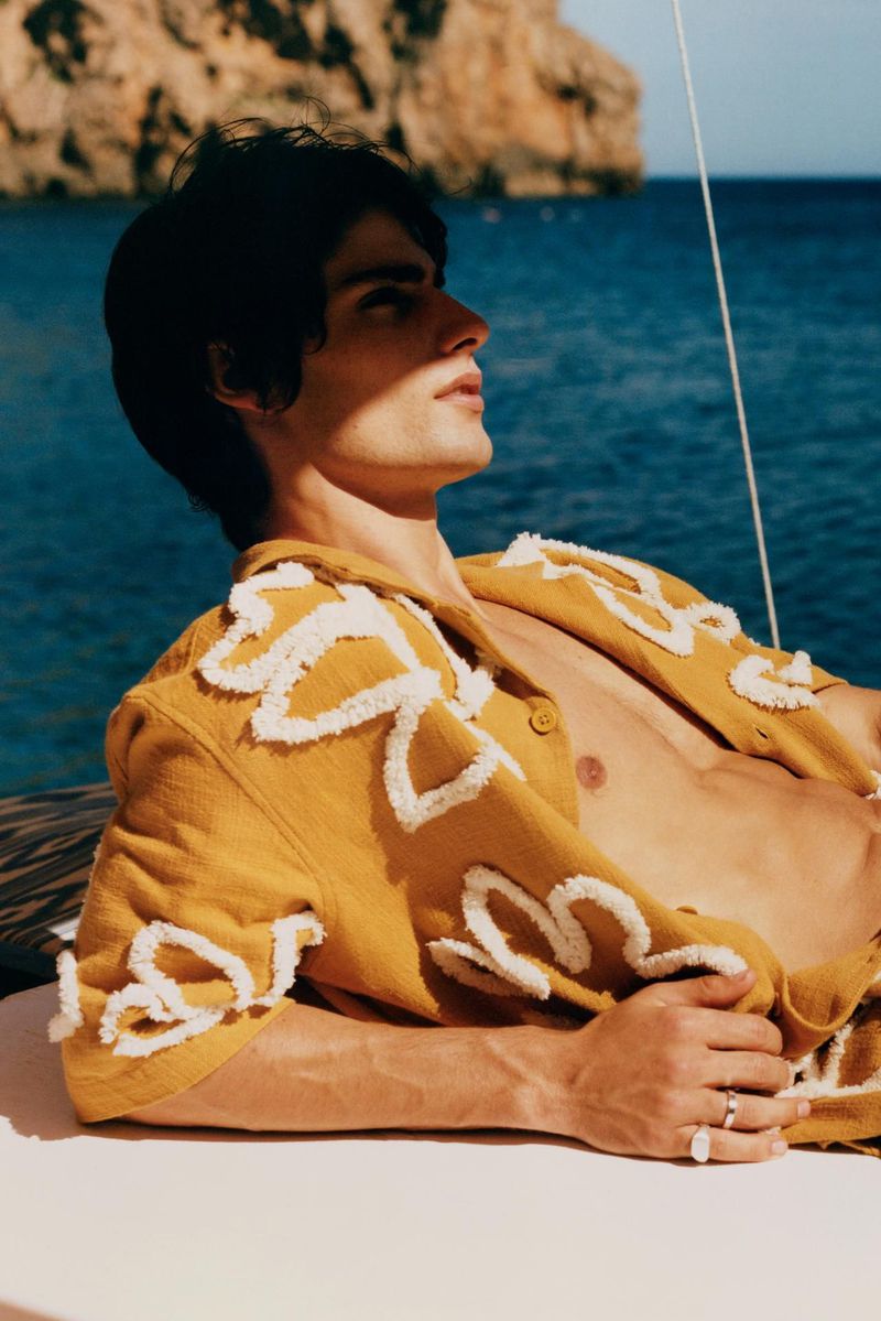 Profile of a light-skinned adult male with dark hair, wearing a patterned yellow shirt, reclining with the ocean and coastal rocks in the background.
