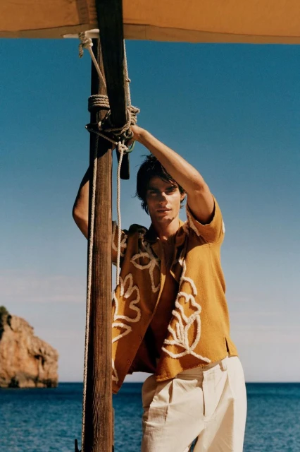 Young adult male poses with a patterned shirt next to a wooden mast on a sunny day by the water near rocky cliffs.