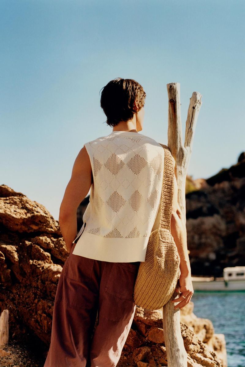 A man stands on a rocky coastline wearing a sleeveless knit top, brown trousers, and a tan shoulder bag, looking out at the sea.