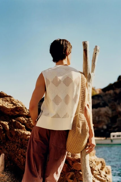 A man stands on a rocky coastline wearing a sleeveless knit top, brown trousers, and a tan shoulder bag, looking out at the sea.