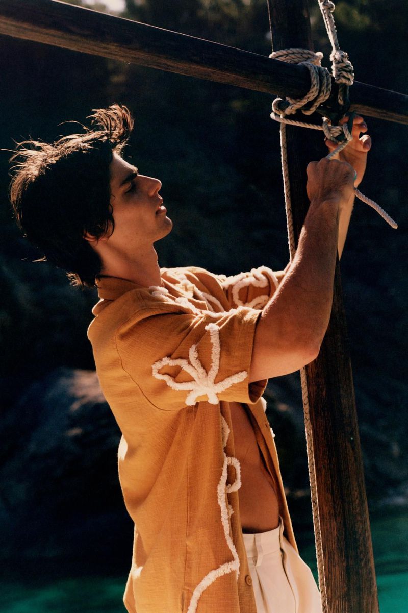 A young adult male is tying a knot with white rope on a wooden post outdoors, wearing a tan short-sleeved shirt and white pants. 