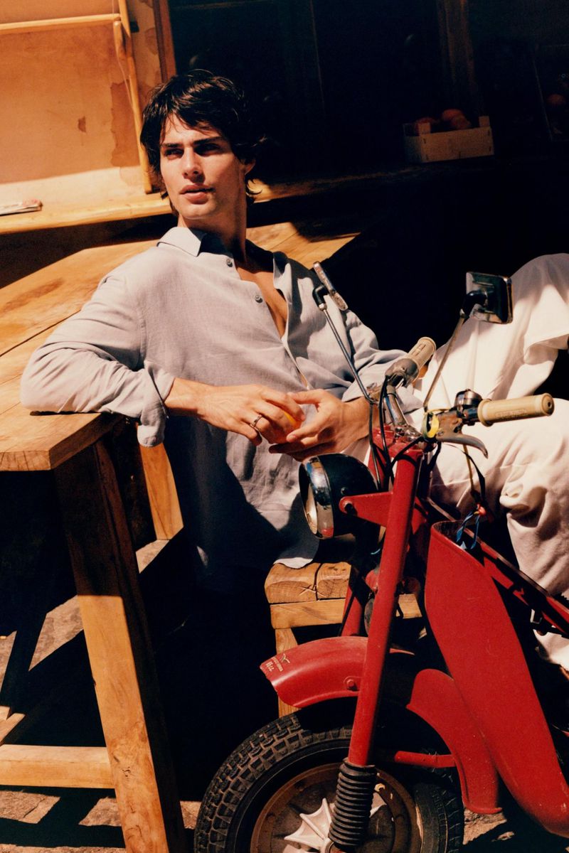 Male model in casual attire poses with a red moped outdoors, showcasing a blend of fashion and automotive style.