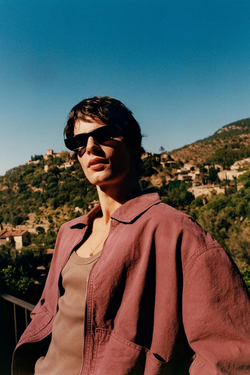 Portrait of a dark-haired adult male model wearing sunglasses and a rose-colored jacket posing outdoors with a hillside town behind.