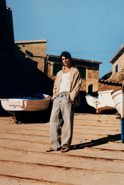 An adult male stands outdoors on a sunny day, wearing jeans and a light jacket, with white boats and stone buildings in the background.