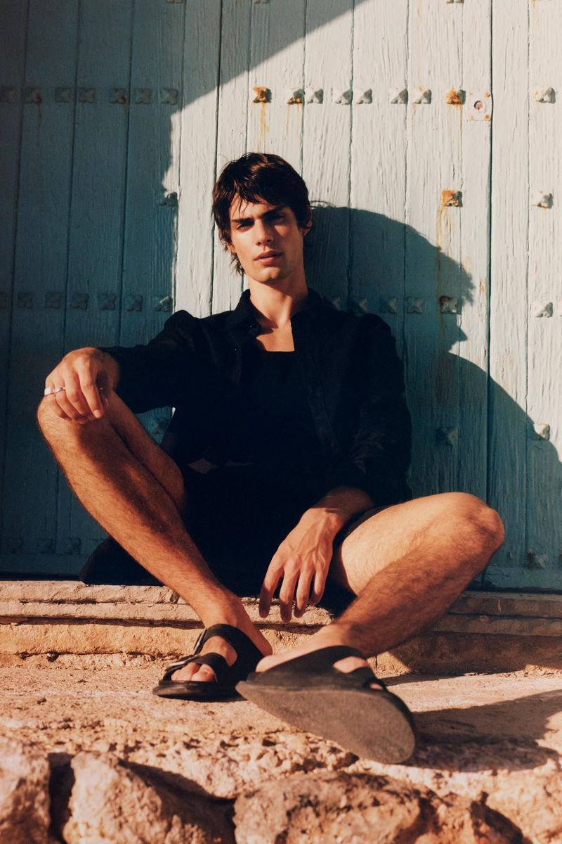 An adult male model sits outdoors in a black jacket, dark shorts, and sandals, in front of a textured blue door with visible sunlight.