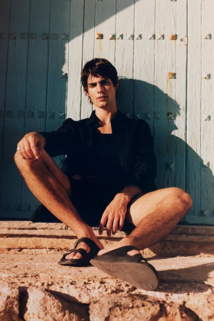 An adult male model sits outdoors in a black jacket, dark shorts, and sandals, in front of a textured blue door with visible sunlight.