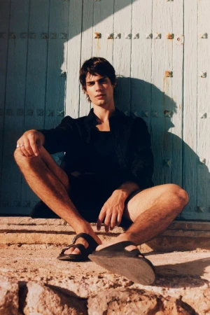 An adult male model sits outdoors in a black jacket, dark shorts, and sandals, in front of a textured blue door with visible sunlight.