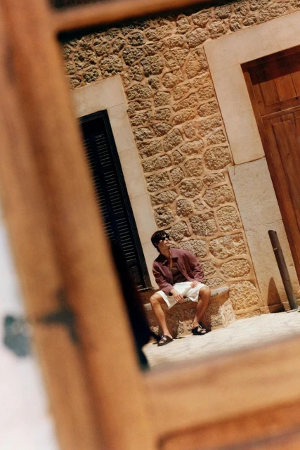 Man sitting on stone ledge in white shorts and sunglasses, reflected in window with building with tan stone brick in background.