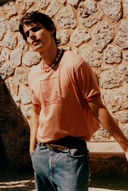 A young man in casual clothing poses against a textured stone wall, wearing a graphic tee, shell necklace, and jeans in natural sunlight.