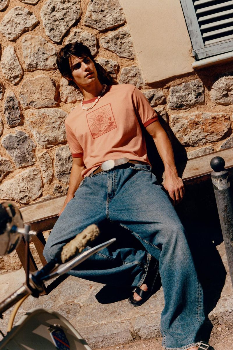 A young man wearing casual clothes sits outside, leaning against a stone wall with a scooter visible; sunlit with cool tones.