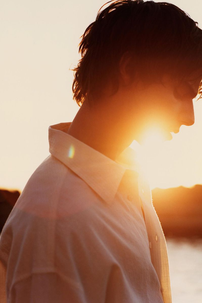 Side profile of a young adult male wearing a collared shirt, backlit by the sun during golden hour, creating lens flare and a warm ambiance.