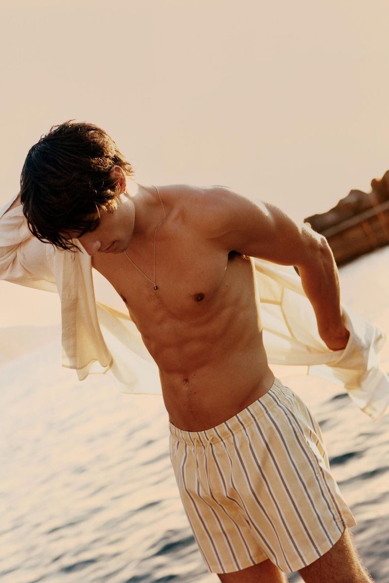 Young man in striped shorts takes off his shirt at a beach location with warm sunlight, showcasing fashion and fitness in an outdoor setting.