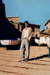 Model standing on a paved waterfront slipway wearing an unbuttoned shirt over a white tank top and baggy light wash jeans.