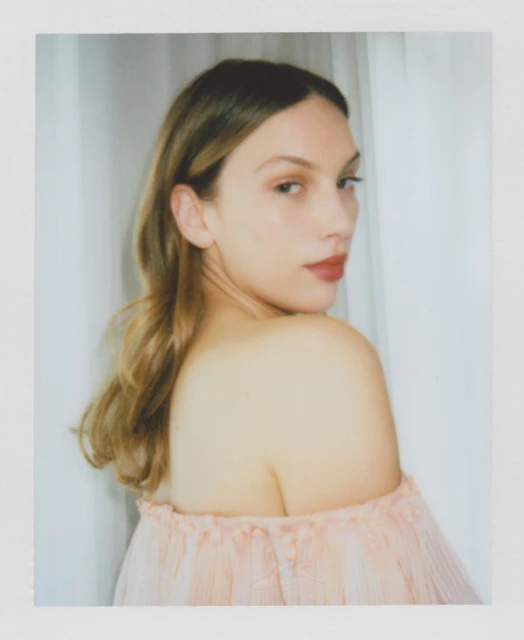 Portrait of a young fair-skinned woman with light brown hair and red lipstick, wearing a pink off-the-shoulder top against a white curtain.