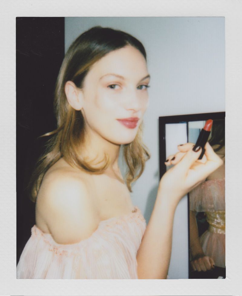 Portrait of a fair-skinned woman applying lipstick with short blonde hair wearing a pink dress indoors with a dark wood door.