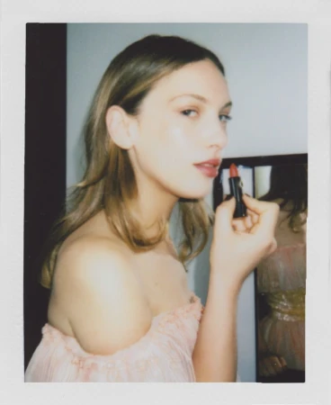 Blonde woman applies lipstick indoors. The close-up portrait captures the beauty routine with natural light illuminating her features.