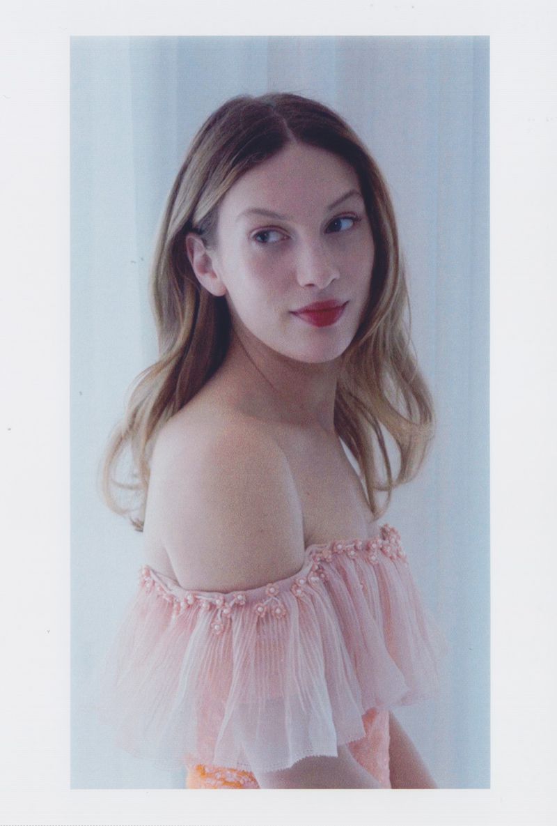 Elegant woman poses indoors wearing an off-the-shoulder pink gown with tulle. She has light brown hair and wears red lipstick.