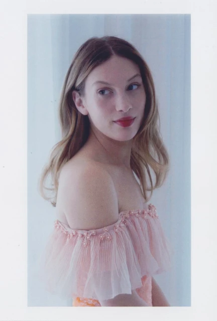 Elegant woman poses indoors wearing an off-the-shoulder pink gown with tulle. She has light brown hair and wears red lipstick.