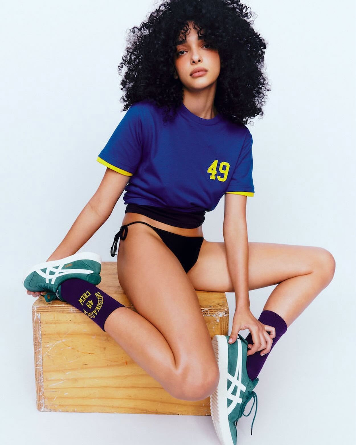 A woman with afro hairstyle poses in a blue t-shirt and black bikini bottoms while sitting on a wooden crate in a light studio.