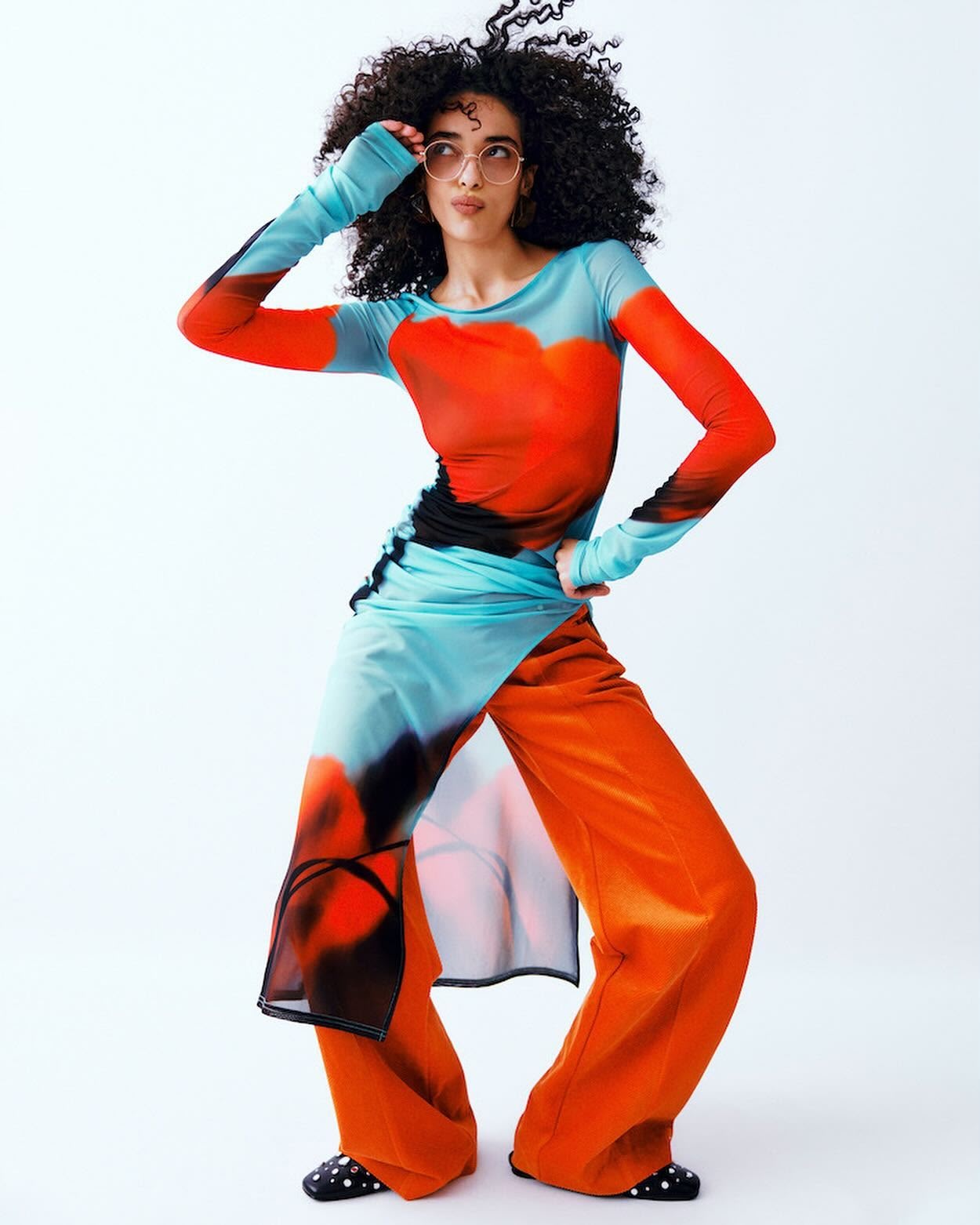 Adult female fashion model poses in a long-sleeved red, black and turquoise print top and orange pants in a white studio.