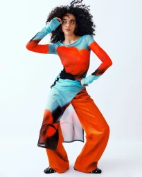Model with voluminous dark curly hair wearing a vibrant orange and blue printed sheer top with matching trousers against white