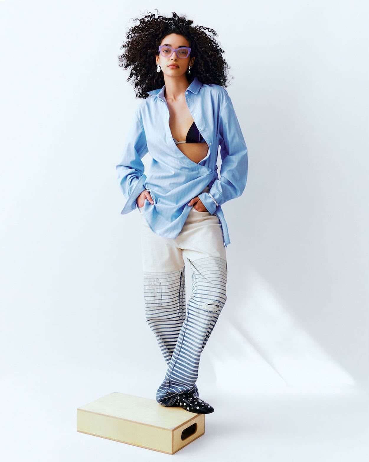 Fashion editorial shot of a young adult female with curly hair posing in a light blue oversized shirt and printed pants on a box in a studio.