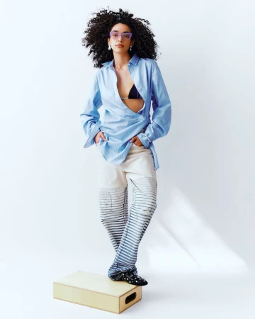 Fashion editorial shot of a young adult female with curly hair posing in a light blue oversized shirt and printed pants on a box in a studio.