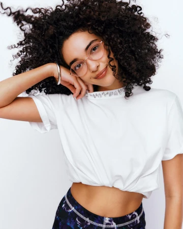 Portrait of a young adult woman with curly dark hair, wearing glasses and a white t-shirt, posing against a white backdrop.