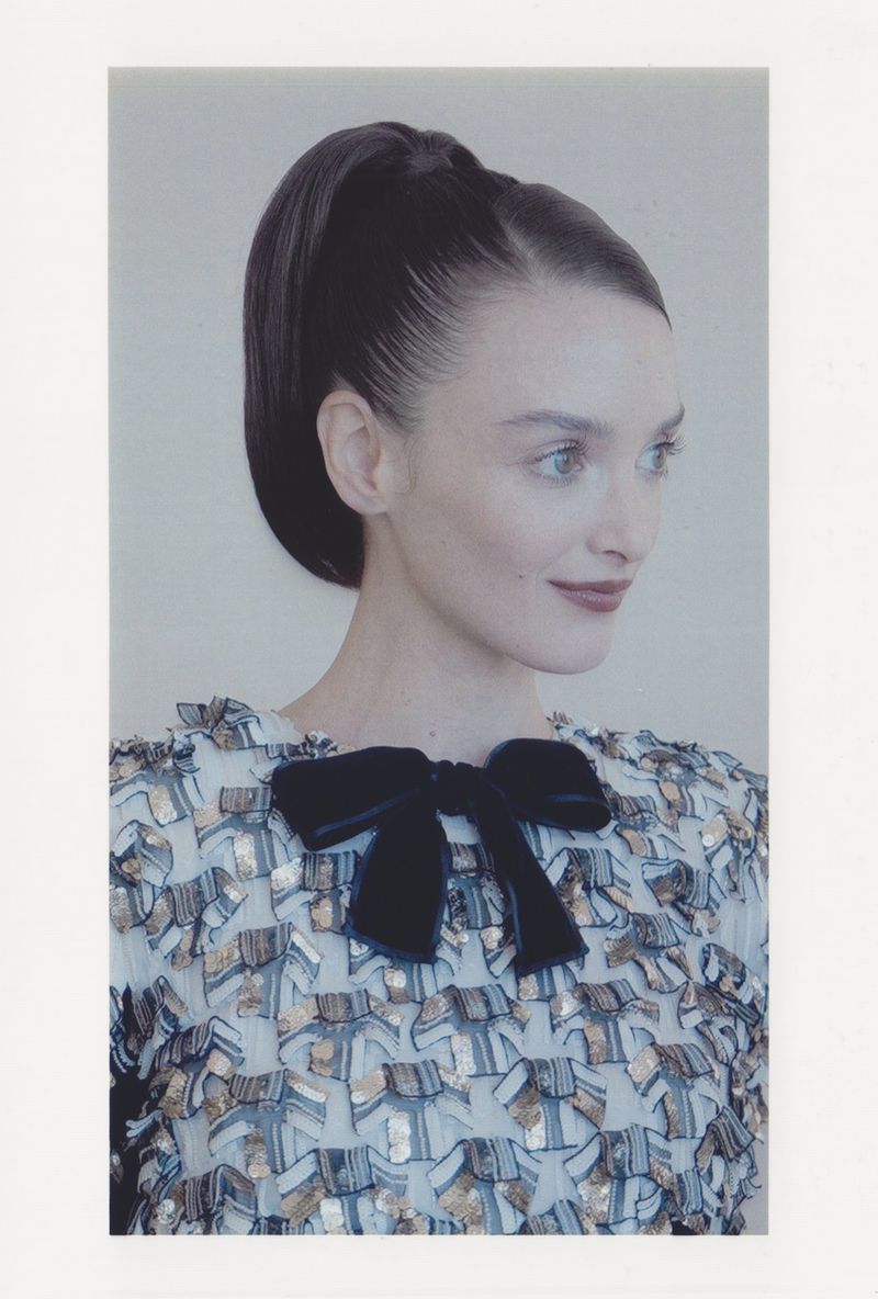 Portrait of an adult female fashion model with dark hair in a high ponytail, wearing a decorative dress and looking off-camera.
