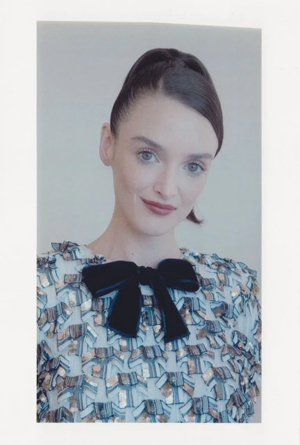 A light-skinned woman with blue eyes and dark hair, wearing a patterned dress with a dark velvet bow, poses indoors against a plain wall.