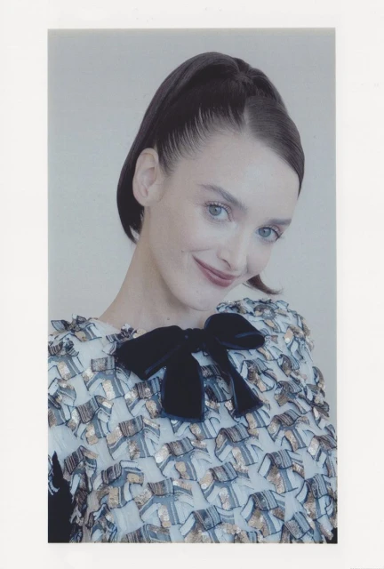 Portrait of an adult woman with dark hair pulled back, wearing a light patterned blouse with a large black velvet bow, smiling.