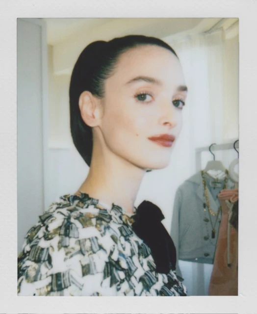Portrait of an adult woman with dark hair pulled back, wearing a patterned top, looking to the side in a soft, indoor setting.