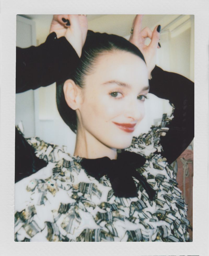 Woman poses with hands near her head in an indoor setting. The image features a close-up shot with a unique polaroid frame aesthetic.