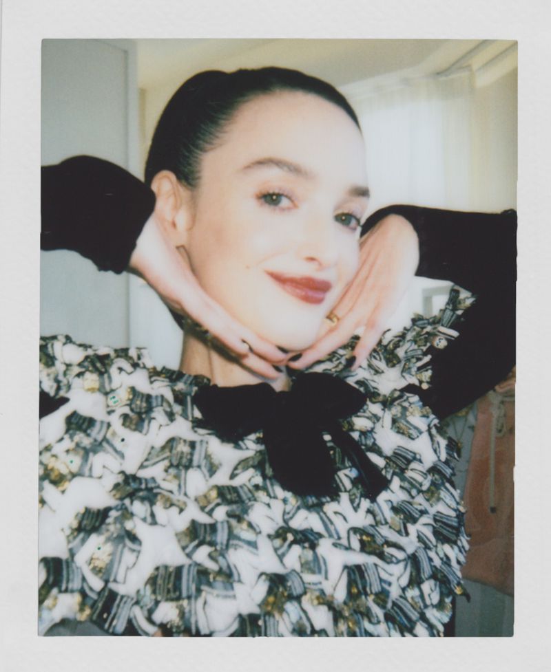 Portrait of a smiling woman with dark hair in a textured top with a bow, posing with hands near her face in an indoor setting.