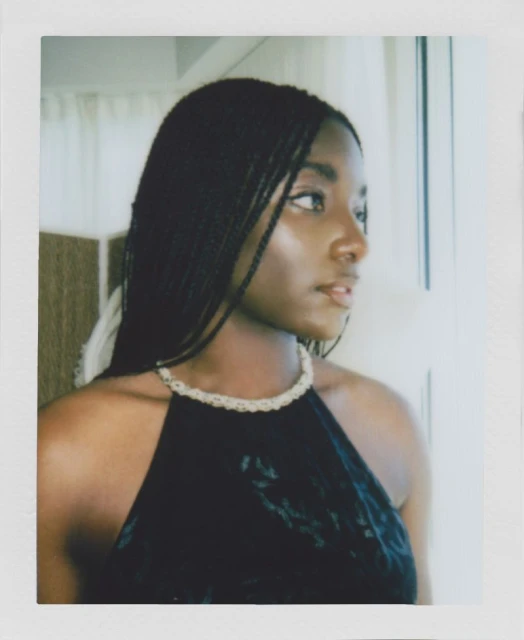 Portrait of a young black woman with braided hair wearing a black halter top and beaded necklace looking out a window with white curtains.