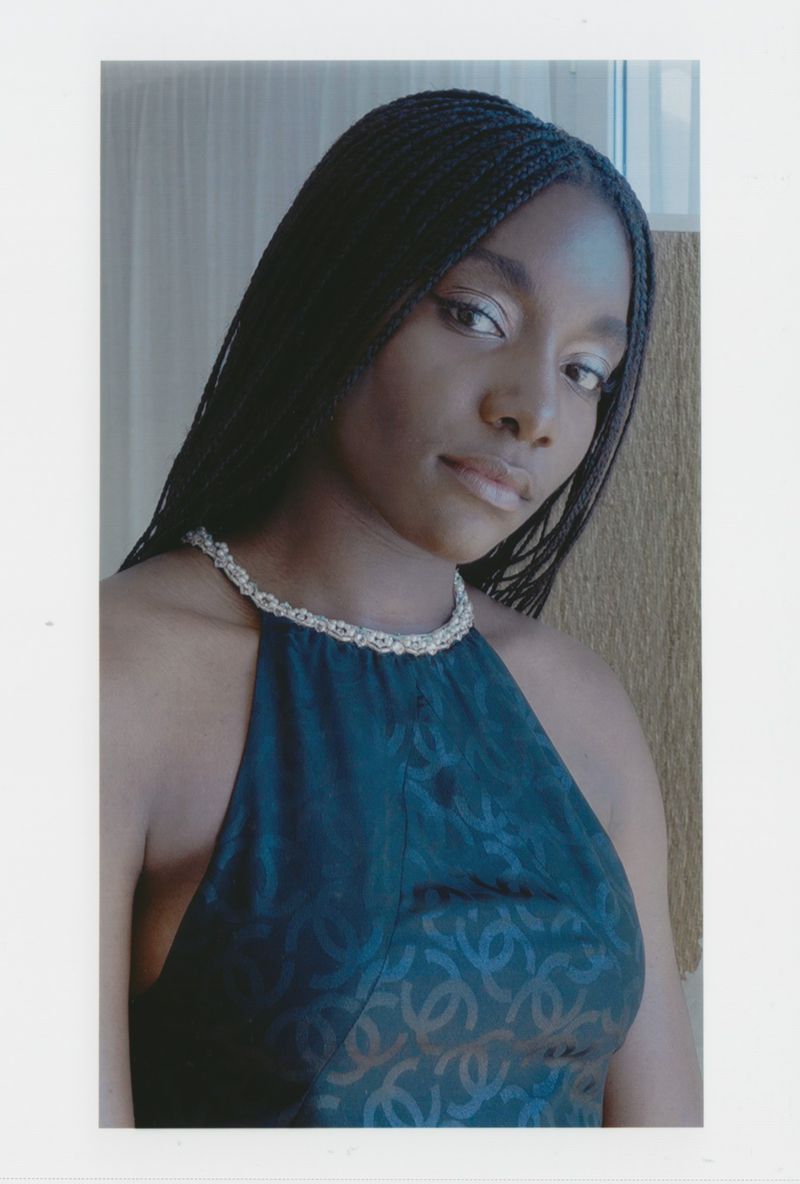 Young woman in a black halter top with braided hair and a pearl necklace posing indoors with a neutral expression and soft lighting.