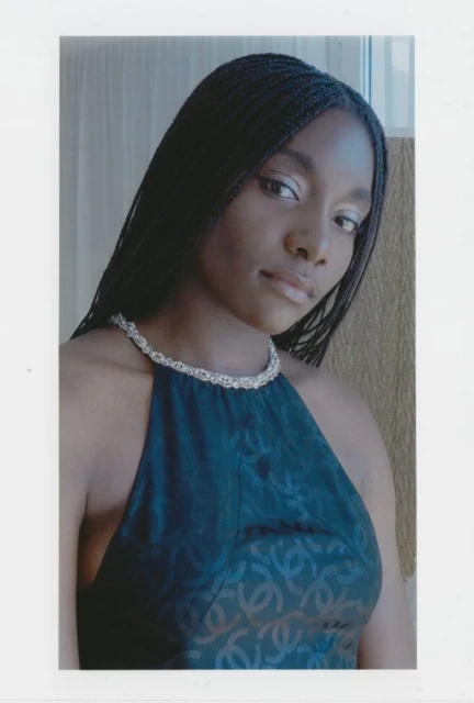 Young woman in a black halter top with braided hair and a pearl necklace posing indoors with a neutral expression and soft lighting.
