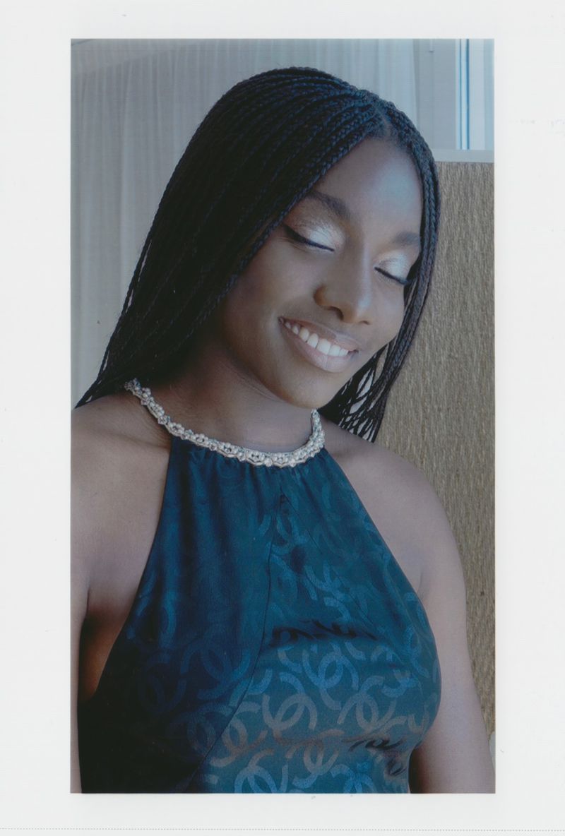 Smiling Black woman with braided hair, wearing a halter dress and silver necklace, eyes closed slightly, indoors with neutral background.