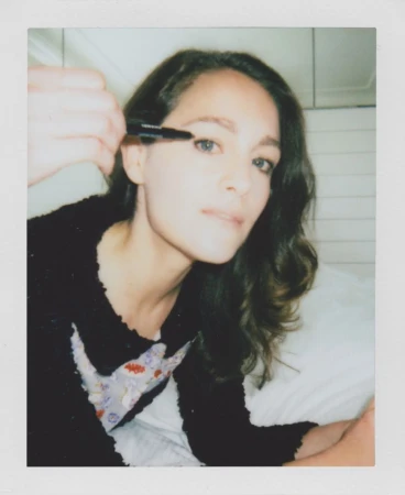 A woman with dark wavy hair applies eyeliner to her top eyelid. She wears a floral-print top with a black cardigan in an indoor setting.