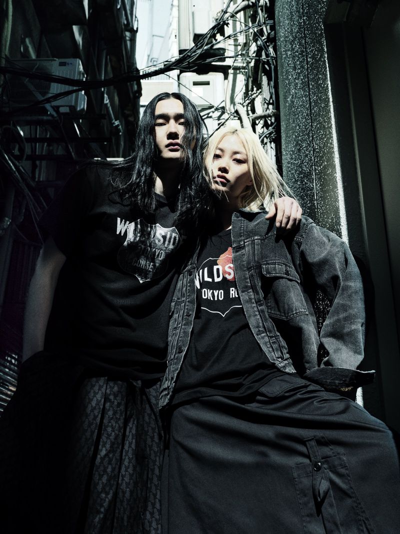 Fashionable couple in dark urban alleyway. Man with long hair and woman in denim jacket pose with edgy style in black clothing.